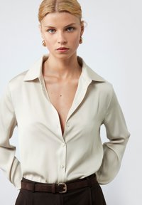 Lichtbeige blouse met een brede kraag en een knoopsluiting aan de voorkant, gemaakt van een gladde stof. Draagt met donkerbruine broek en een bijpassende riem.