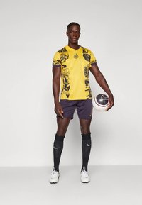 Žltý futbalový dres s čiernymi geometrickými vzormi, v kombinácii s tmavými šortkami, čiernymi podkolienkami a bielymi futbalovými kopačkami. Drží loptu.