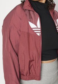 Vínová větrovka s mírným leskem, zdobená bílými logy Adidas, se zipem na zapínání a elastickým lemem a manžetami.