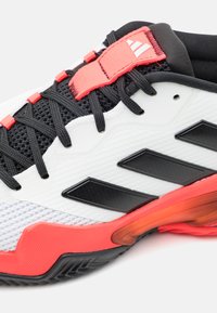 Vit och svart sportskor med röda accenter; har en texturerad yta, meshdetaljer och framträdande svarta ränder på sidan.