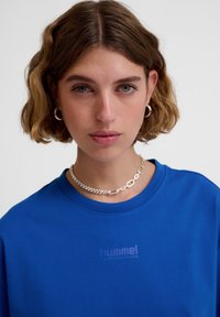 Blå bomullströja med rund halsringning och korta ärmar, med en subtil präglad logotypdetalj i mitten. Silverkedjehalsband som accentuerar.