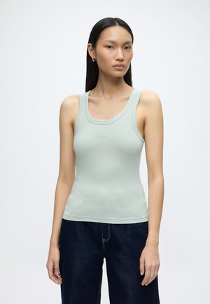 Jeune femme aux longs cheveux noirs et lisses, portant un débardeur vert clair sans manches et un pantalon bleu foncé sur un fond blanc uni.