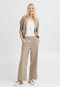 Blazer beige à manches longues, associé à un pantalon large beige et un top blanc. Le mannequin se tient avec les mains dans les poches. Texture de tissu lisse.