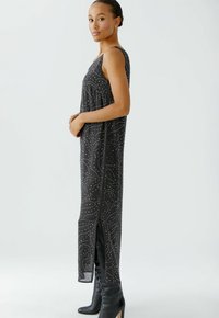 Robe noire sans manches au fit ample, avec un motif texturé de pois blancs et des fentes sur les côtés. Portée avec des bottes noires montant jusqu'aux genoux.