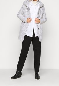 Cappotto grigio texturizzato con cappuccio, chiusura con bottoni e cerniera, indossato sopra una camicia bianca e pantaloni neri. Stivaletti alla caviglia neri con motivo completano il look.