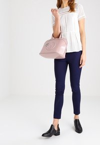 Różowa, lakierowana torebka o zaokrąglonym kształcie, z górnymi uchwytami i wyraźnym detalem logo. Noszona z białym dzianinowym topem i granatowymi spodniami.