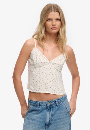 Jeune femme aux longs cheveux blonds portant un crop top blanc à fines bretelles à motifs floraux et un jean en denim bleu sur un fond uni.