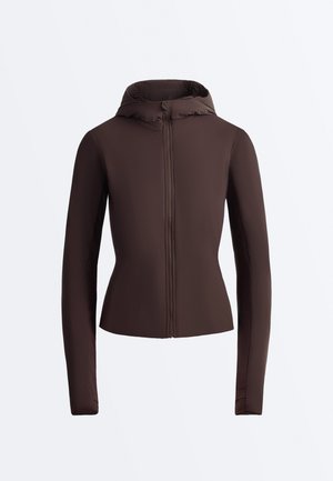 Mørkebrunt tætsiddende langærmet jakke med lynlås foran og hætte, vist på en ensfarvet lysegrå baggrund.