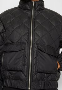 Czarna pikowana kurtka puchowa z wysokim kołnierzem, srebrnym zamkiem i dwiema przednimi kieszeniami z klapką. Materiał jest gładki i ma lekką teksturę.