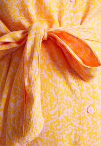 Um tecido amarelo e rosa com padrão apresenta uma cintura amarrada com um nó. Inclui botões rosa na frente. A textura parece leve e tecida.