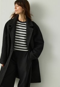 Woman wearing a textured black coat, black wide-leg pants, and a black-and-white striped top, looking to the left against a plain wall.