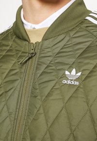 Olivgrüne, gesteppte Bomberjacke mit geripptem Kragen, Reißverschluss und weißem Adidas-Logo. Verfügt über ein strukturiertes Rautenmuster und einen Reißverschlussanhänger.