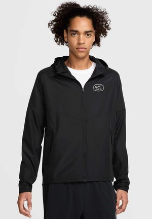 Chaqueta negra ligera con cierre frontal, capucha y bolsillos laterales. Presenta un logo reflectante de Nike Air y un acabado suave y texturizado.