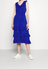 Blå lager-på-lager klänning med volangaxlar och knappdetaljer, med en dragsko i midjan. Bärs med vita sneakers. Texturerad tyg.