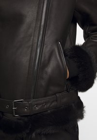 Svart läderjacka med dragkedja, sidfickor och en texturerad faux fur-kant längs nederkant och ärmender. Bälte i midjan.