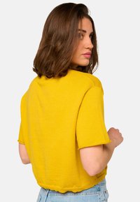 Femme aux cheveux châtains mi-longs portant un haut jaune à manches courtes et un jean bleu clair, légèrement de profil sur un fond blanc.