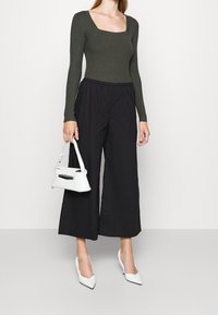 Frau trägt ein olivgrünes, ribbed langärmliges Oberteil, schwarze weit geschnittene 3/4-Hosen, weiße spitze Absatzschuhe und hält eine kleine weiße Handtasche.