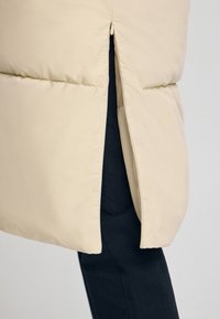 Giacca puffer beige con una ventilazione laterale con zip, caratterizzata da una texture liscia e un motivo a cuciture orizzontali. Tessuto scuro visibile sotto.