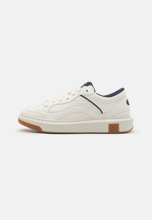 Basket blanche basse avec doublure marine, lacets blancs, panneaux latéraux texturés et semelle en caoutchouc marron avec le logo "A|X" près du talon.