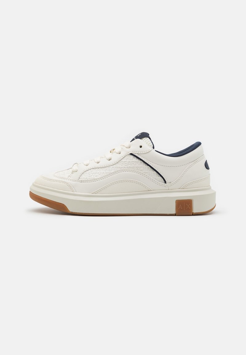 Basket blanche basse avec doublure marine, lacets blancs, panneaux latéraux texturés et semelle en caoutchouc marron avec le logo "A|X" près du talon.