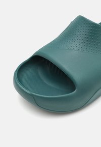 Zöld csúszásmentes szandál rugalmas gumiból, texturált felső résszel, nyitott orral és finom perforációkkal a szellőzés érdekében.