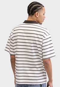 Polo a maniche corte di colore bianco con righe orizzontali marroni, colletto a coste marrone, tessuto in cotone, vestibilità comoda e spacchi laterali.