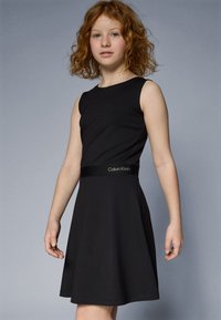 Melnā bezpiedurkņu kleita ar izplūdušu svārku, auduma josta ar Calvin Klein logotipu. Gluda tekstūra un klasiskā silueta.
