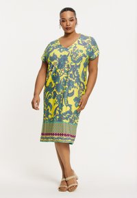 Kurzärmeliges gelbes Kleid mit blauen Blumenmustern, V-Ausschnitt, knielang und einem Saum mit grünem und lila Muster. Sandalen runden den Look ab.