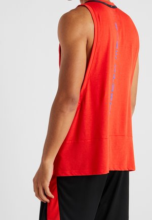 Atlet iført ærmeløs rød Nike Sport-top med vertikalt logo på ryggen og sorte shorts foran en ensfarvet baggrund.