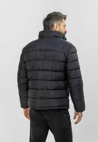 Black puffer jacket with a high collar, featuring horizontal quilted sections. Smooth, shiny fabric with a relaxed fit. Worn with dark jeans.