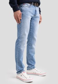 Mees, kes kannab helerohelisi teksapükse, pruuni vööd, mereväe sinist pusa ja valgeid madalaid tossusid, seisab ühevärvilise tausta ees.