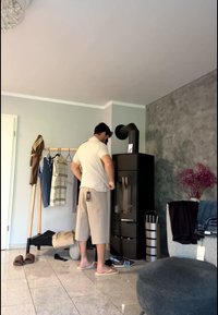 Άνδρας ντυμένος με μπεζ ρούχα στέκεται κοντά σε μαύρη ξύλινη σόμπα σε φωτεινό δωμάτιο με κρεμάστρα για ρούχα, παντόφλες και γκρι πολυθρόνα με διπλωμένα ρούχα.