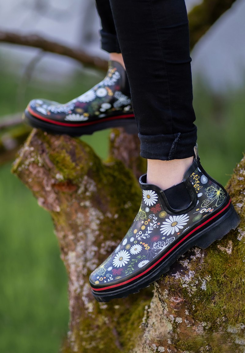 Schwarze Gummistiefel knöchelhoch mit einem floralen Muster in Weiß, Grün und Rot, ausgestattet mit einer strukturierten Sohle und einem roten Akzent am Boden.