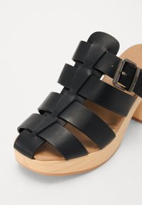 Fekete bőr slip-on szandál szőtt pántokkal, fa talpbetéttel és állítható fémcsatként. Simává varázsolt felület és nyitott orrdesign.