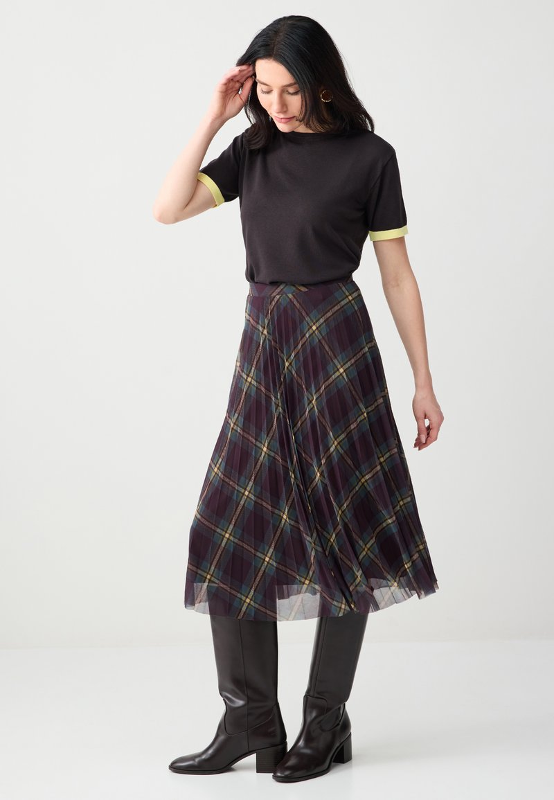 Femme portant un t-shirt noir à manches courtes avec des poignets jaunes, une jupe midi plissée à carreaux violets, et des bottes noires montantes, debout devant un fond blanc.