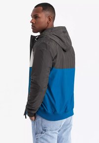 Giacca color block con cappuccio grigio, sezione superiore bianca e parte inferiore blu. Realizzata in materiale leggero e liscio con polsini elasticizzati.