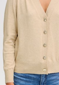 Cardigan beige en matériau doux, avec un col en V, des boutons dorés et des poignets côtelés, associé à un jean en denim bleu.