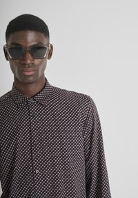 Donker gemusterde shirt met een geometrisch ontwerp in rood en wit, met een kraag en knopen aan de voorkant. Zwarte zonnebril maakt de look compleet.