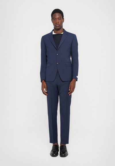 Costume bleu marine avec un blazer ajusté et un pantalon droit. Le blazer est doté de deux boutons, de poches latérales et d'une texture lisse. Chaussures noires.