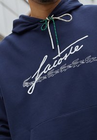 Tmavě modrá bavlněná mikina s kapucí s bílým nápisem ve skriptovém stylu a vícebarevnými šňůrkami na stažení. Design zahrnuje malé vyšívané krokodýly.