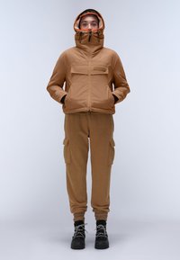 Giacca isolata marrone con colletto alto, cappuccio foderato in arancione, tasche con zip, abbinata a pantaloni cargo beige chiaro e stivali neri con lacci.
