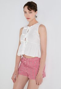 Top bianco senza maniche con orlo arricciato e dettagli a fiocco decorativi; abbinato a pantaloni corti a quadri rosso e bianco.