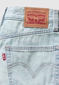 Světle modré džíny z denimu s hnědou koženou nášivkou nesoucí nápis "Levi Strauss & Co.". Náplast má červený text a motiv koní, s klasickým šitím.