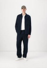 Navy button-up shirt, white T-shirt, loose navy pants, and white sneakers. Gray cap with embroidered detail. Plain, smooth texture.