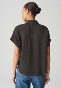Blusa nera a maniche corte con superficie texturizzata, colletto e vestibilità morbida. Il retro presenta un pannello liscio e un orlo leggermente curvo.