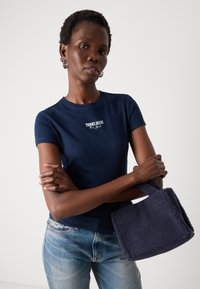 Tamsiai mėlynas trumpomis rankovėmis marškinėliai su balta "TOMMY JEANS New York" užrašu, derinami su šviesiai mėlynomis džinsomis ir tamsiai mėlyna puria rankine.