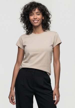 Frau mit lockigem Haar, die lächelt, trägt ein beigefarbenes kurzärmeliges Hemd und schwarze, hoch taillierte Hose, steht vor einfarbigem Hintergrund.