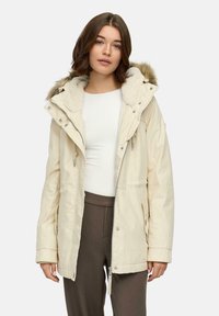 Beige parka-jack met een gevoerde capuchon van nepbont, voorzien van een front-rits, aantrekkoorden en twee zijzakken. Glad stof en relaxed fit.