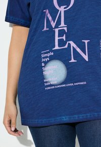 Modrá košile s velkými růžovými písmeny "OMEN" a menším textem "Jednoduché radosti & Letní noc, Severovýchodní strana Západu, Navždy milovník slunce, Štěstí" na přední straně.