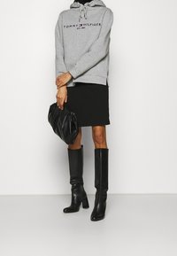 Person som bär en grå Tommy Hilfiger hoodie, svart kjol, svarta knähöga stövlar och håller en avslappnad svart läderklutch.